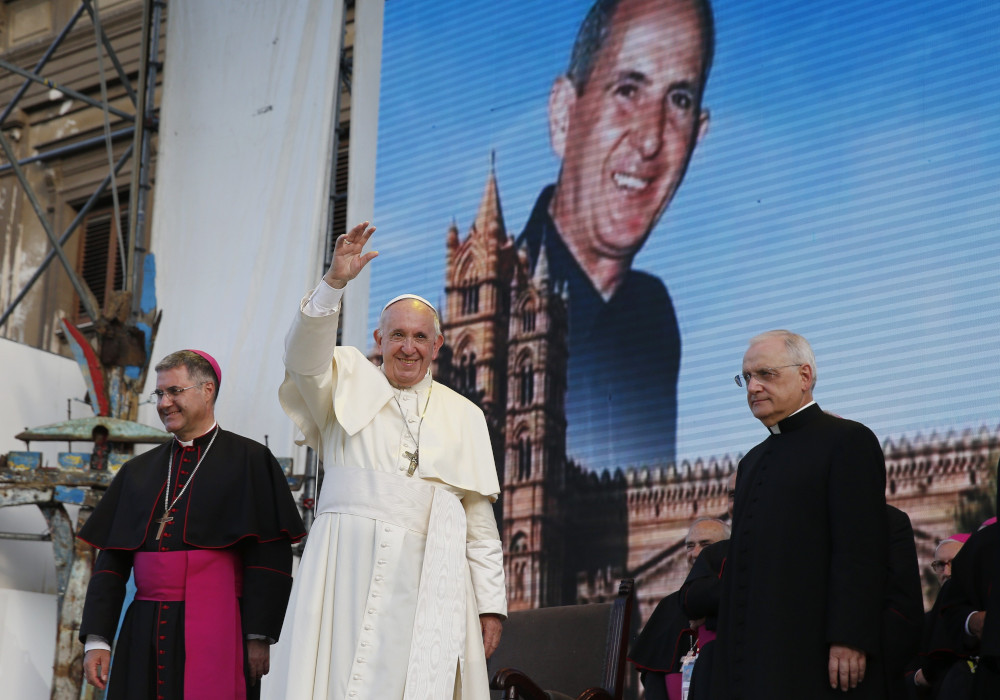 Pope Francis raises his hand to wave while standing in front of a large image of a white priest