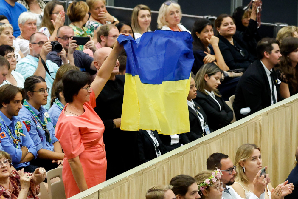 People in a sitting crowd stand with a Ukrainian flag, including a Brown woman with an orange dress