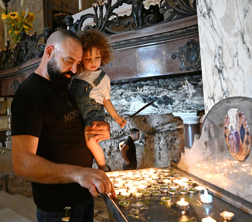 A bearded brown man holds a child in one arm and adjusts a candle with the other hand, while in a church