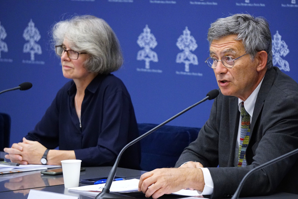 An older white man in a suit speaks into a microphone while seated at a table where an older white woman is present