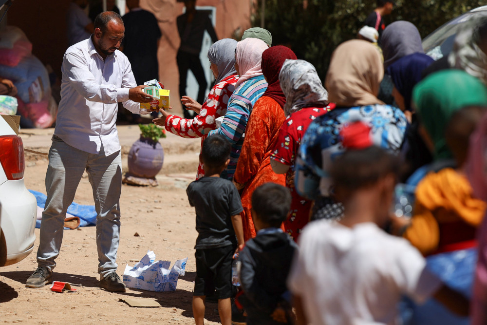 A man hands small boxes to a line of women dressed in colorful dresses and head scarves