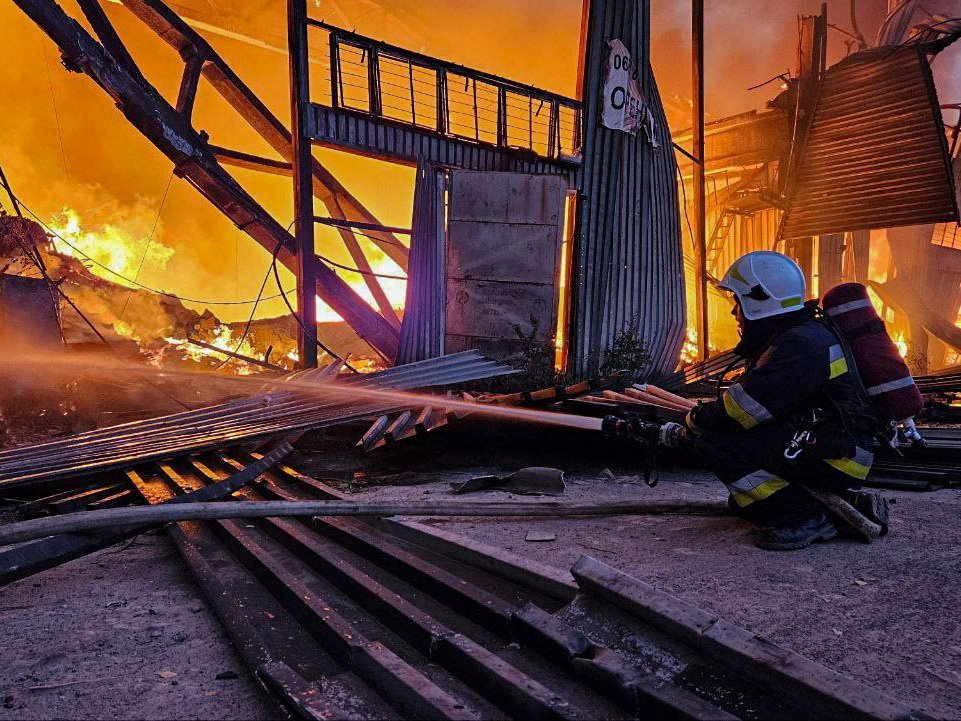 A person in a white helmet sprays water into a landscape full of fire and a burned out building