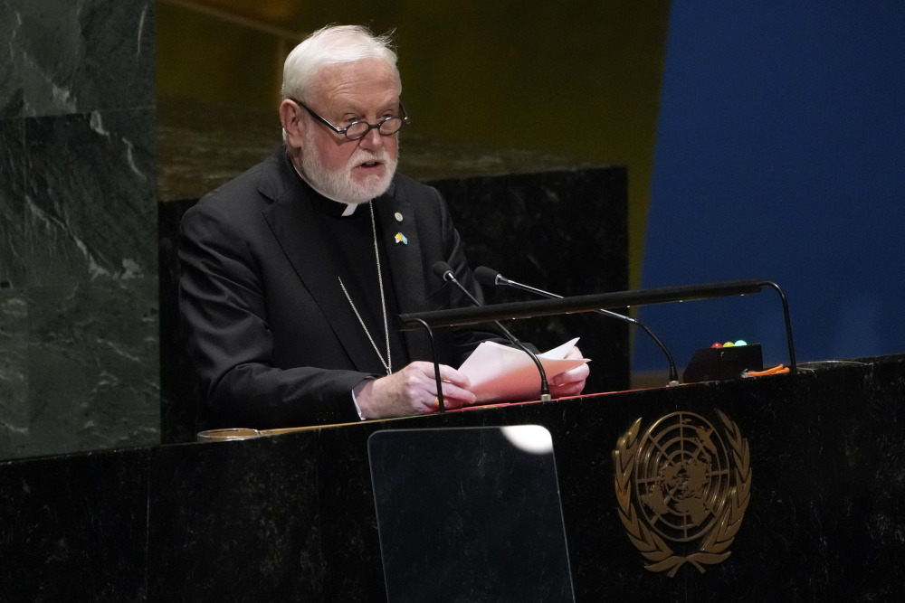 An older white man with white hair and a beard and glasses wearing a clerical collar speaks into a microphone