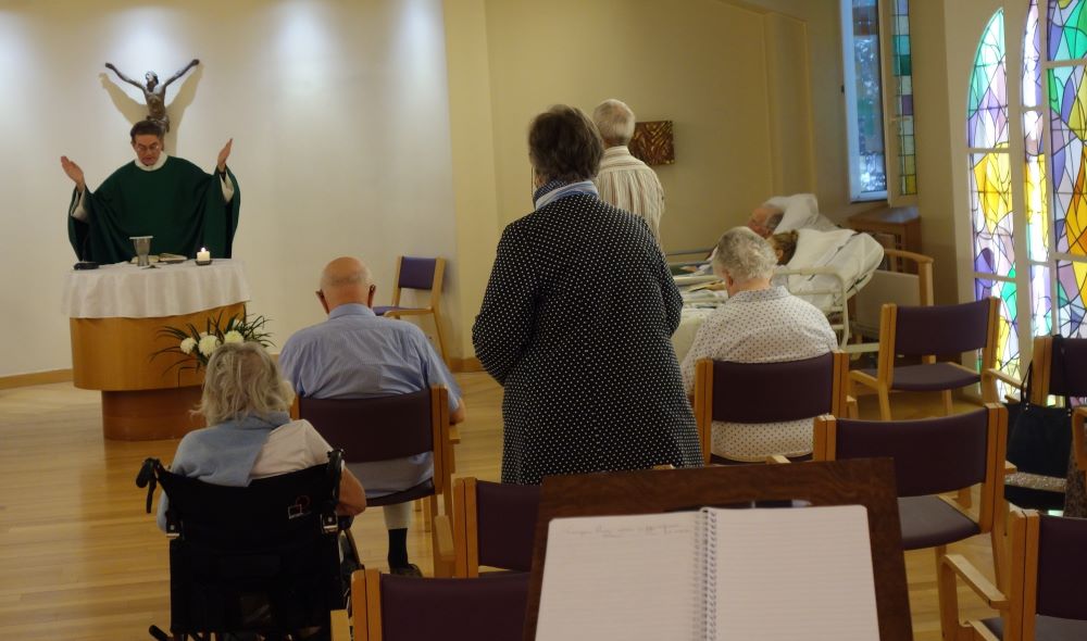 Priest at lectern, leading <ass to people sitting in chars.