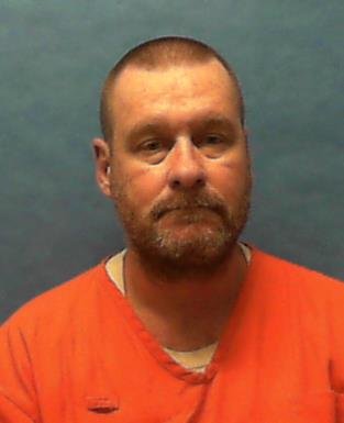 A white man with a brown buzz cut and beard wearing an orange prison shirt looks at the camera