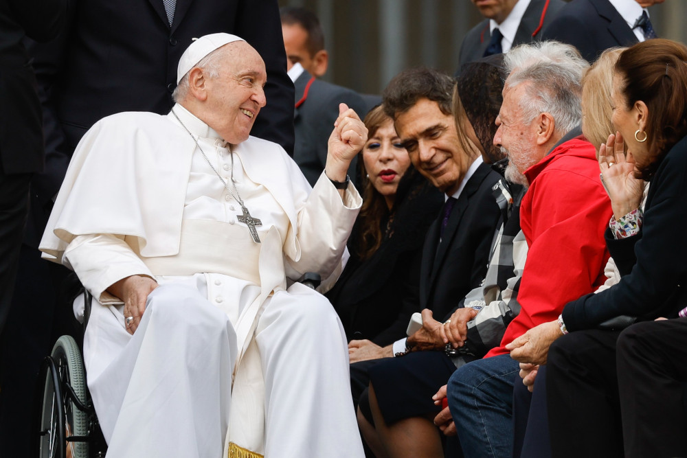 Pope Francis sits in his wheelchair and leans toward and smiles at crowd members as he is wheeled by them