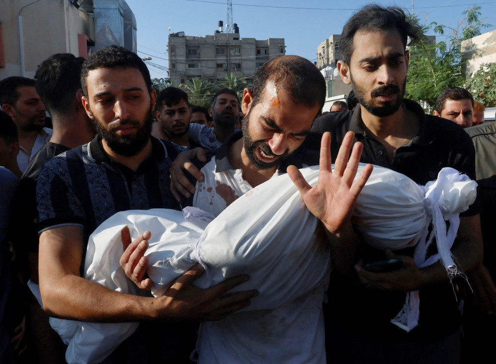 A man hangs his head and cries out, holding a body wrapped in a white sheet, while two men on either side of him hold him up