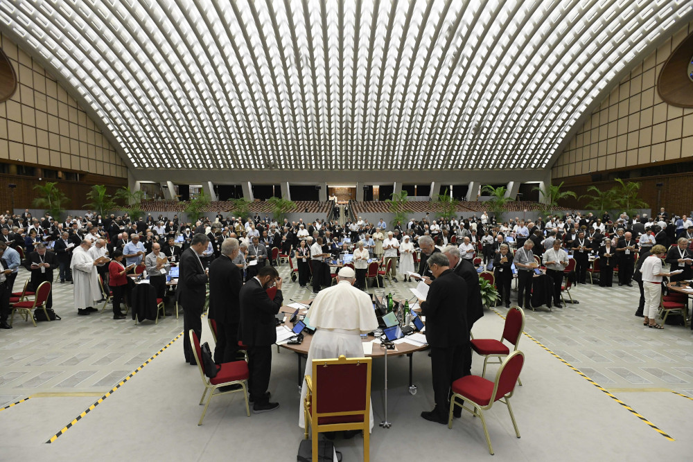 Many people stand around many round tables in a domed hall. Pope Francis is visible at a table in the front.