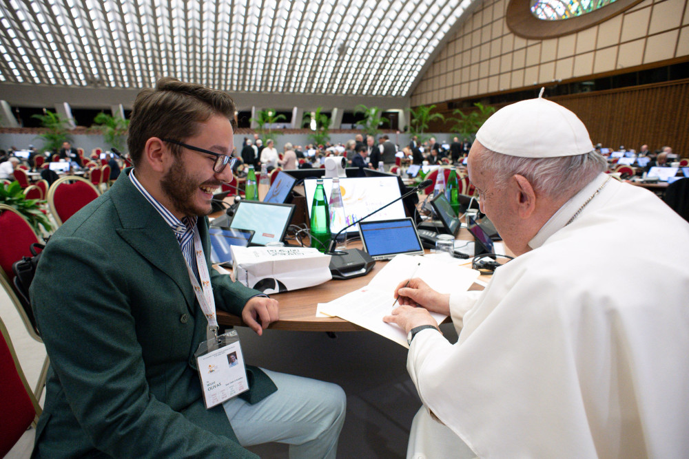 A young man with brown hair wearing a suit smiles at Pope Francis as he signs a document