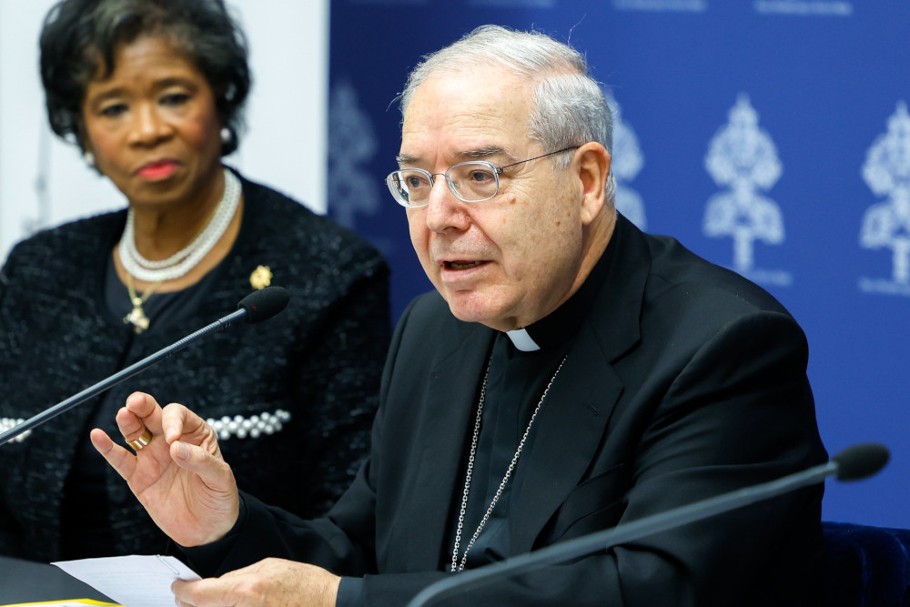 An older white man with glasses wearing a clerical collar speaks into a microphone while a professionally dressed older Black woman looks on