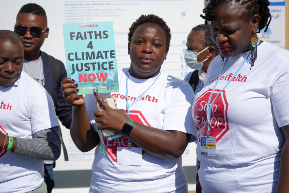 A woman stands with two others in white T-shirts, holding a sign that reads, "Faiths 4 Climate Justice Now."