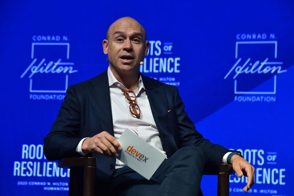 Man sits, looking forward, holding papers, and speaks.