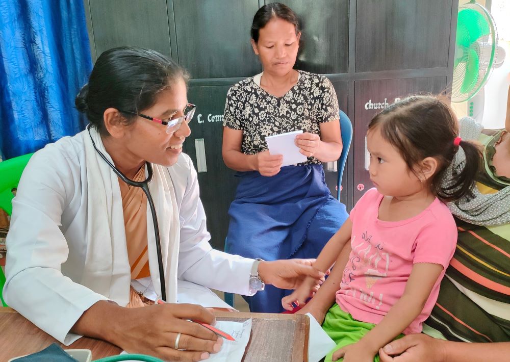 Woman wearing doctor's jacket and with stethoscope talks to little girl and touches her arm.