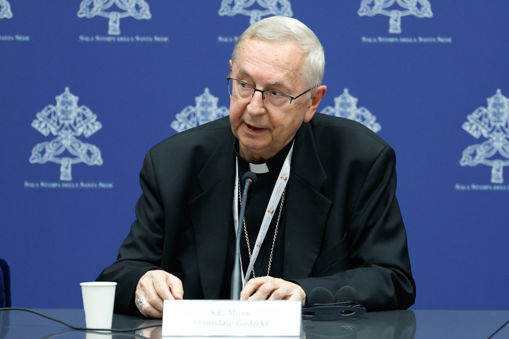 An older white man wearing glasses and a clerical collar speaks from behind a table and in front of a blue background with emblems 