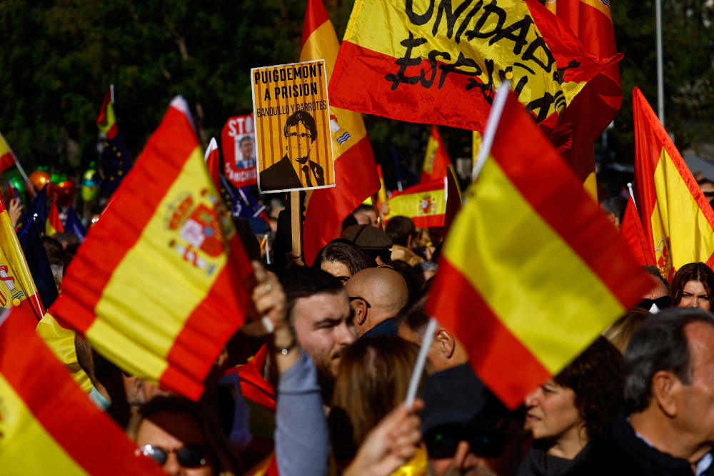 A crowd of people hold up Spanish flags in the dark. One flag has writing on it. 