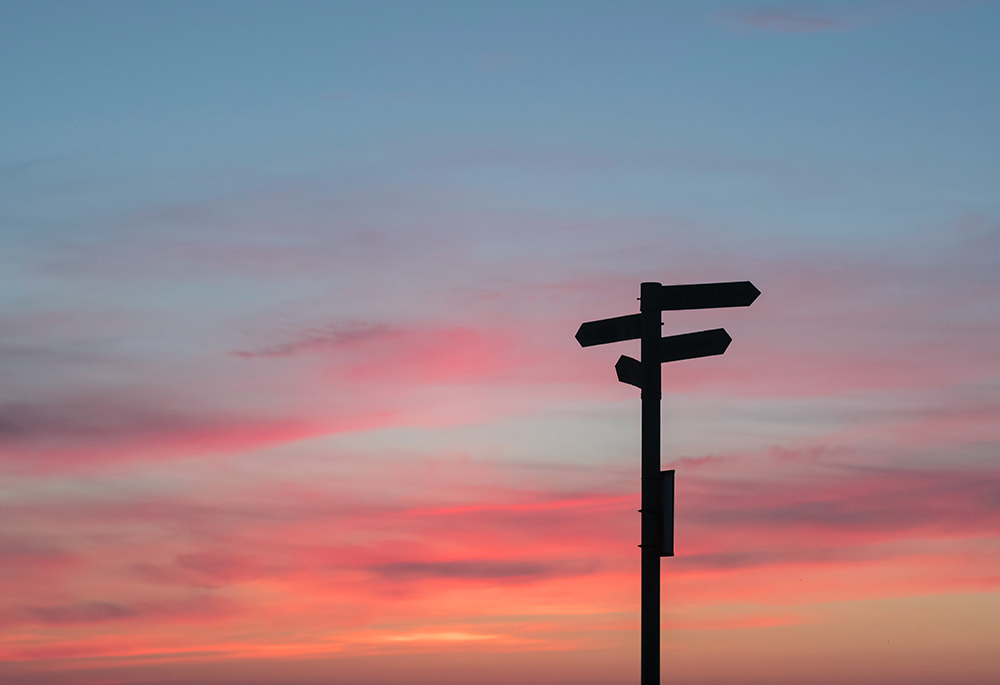 A crossroads street sign is pictured in shadow against a colorful sunset. (Unsplash/Javier Allegue Barros)