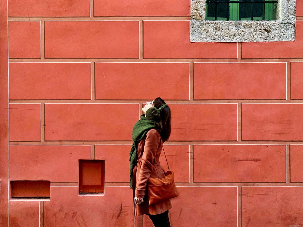 Woman wearing coat and scarf stands outside building with brick walls and looks upward. 