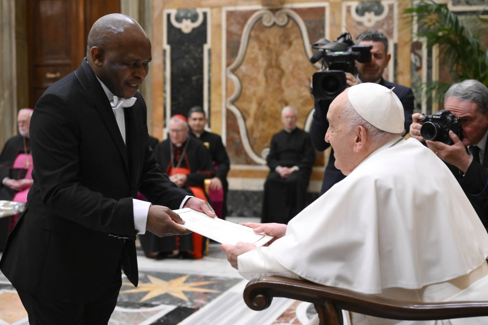 A Black man standing and wearing a white-tie tuxedo hands a seated Pope Francis a piece of paper