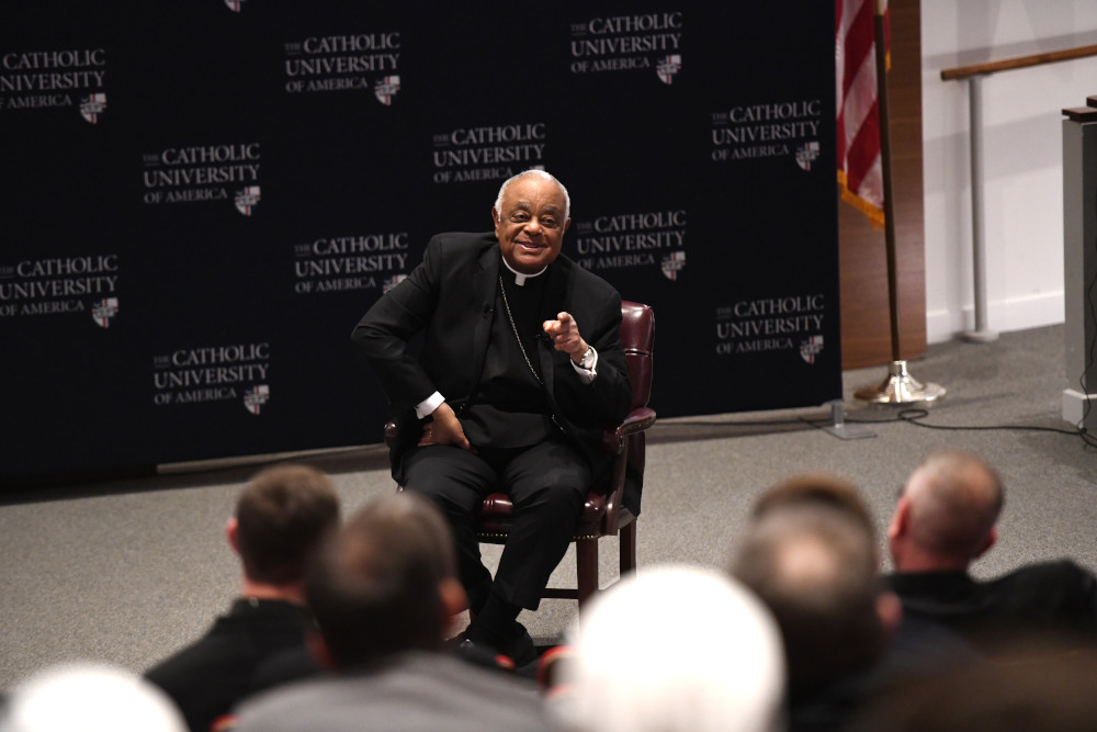 An older Black man wearing a clerical collar and black jacket sits in front of an audience