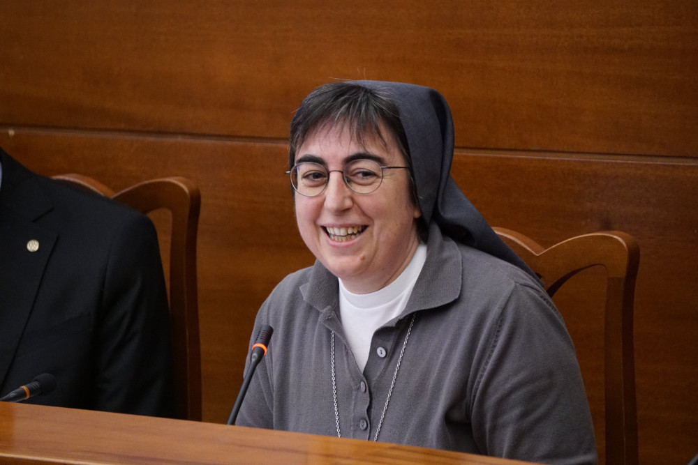 A woman religious wearing glasses and a gray habit sits at a table and speaks into a microphone