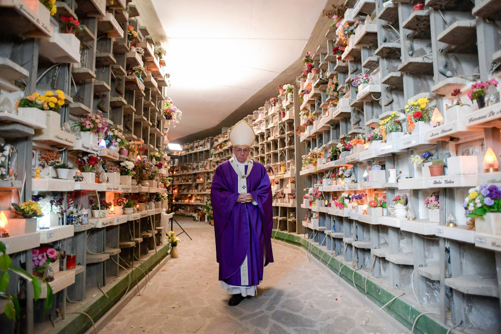 Pope Francis, wearing purple vestments, walks down a hallway with stone shelves and flowers on his sides