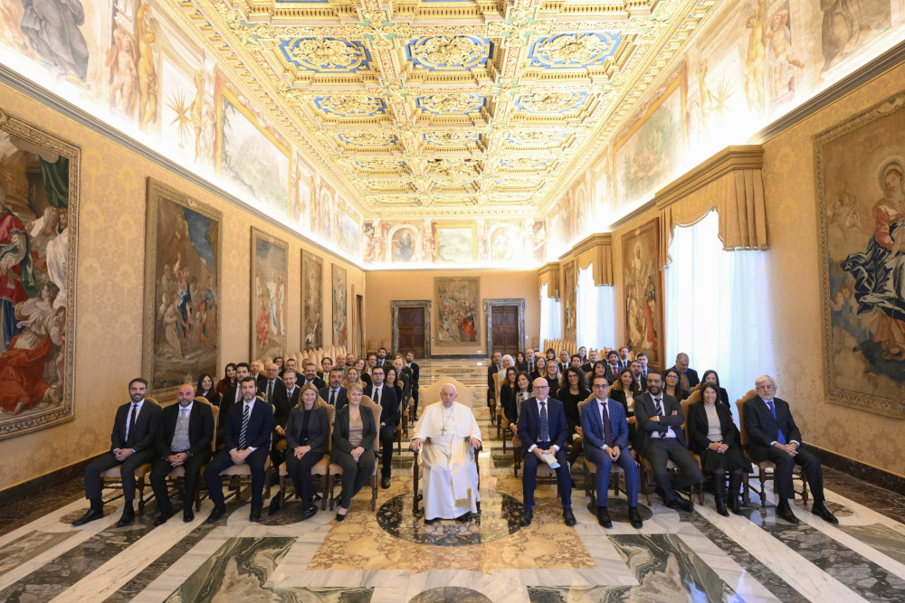 Pope Francis sits in a wheelchair between two sections of seated people in business attire