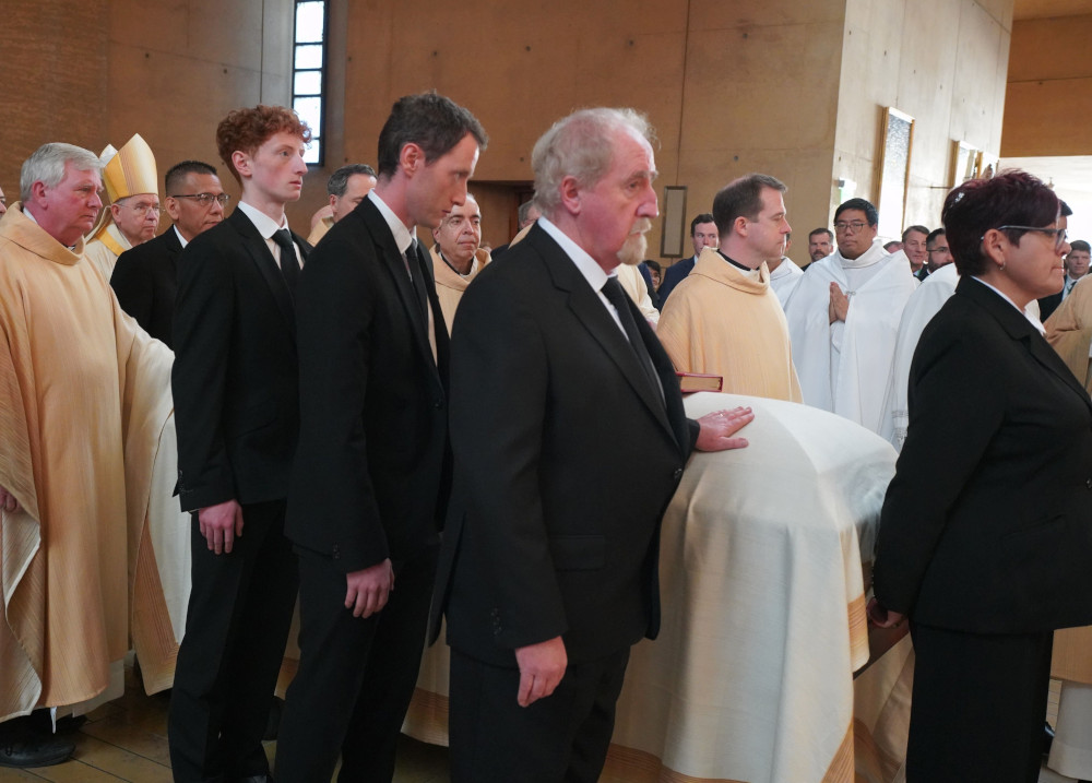 Men in suits and vestments surround a casket draped in a white sheet 