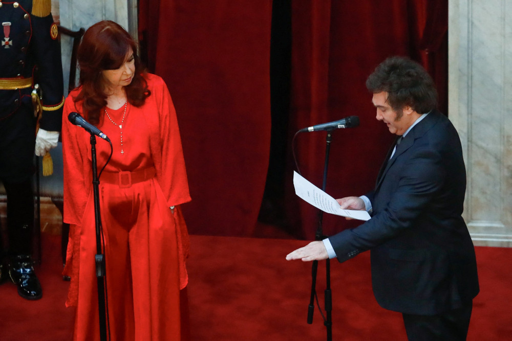 A white man in a suit reads from a paper with his other palm face down while standing next to a woman in a red dress