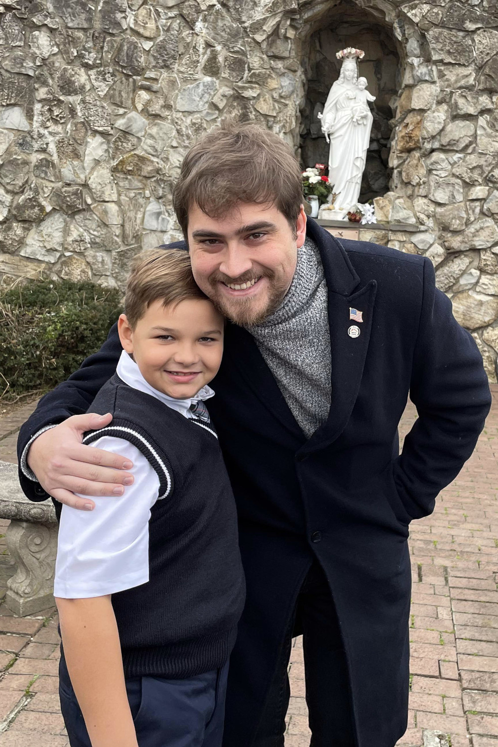 A man wearing a turtleneck and a jackets leans down to pose with a boy in a sweater vest. A statue of Mary is visible in the background.