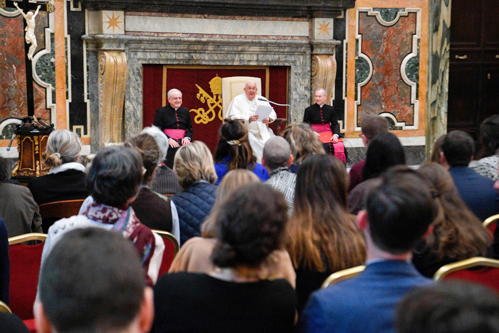 Pope Francis sits in front of a crowd, holding a piece of paper and speaking into a microphone