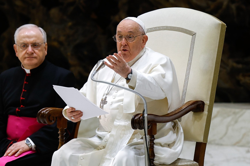 Pope Francis speaks and gestures while sitting in his white chair. A man wearing a bishop's cassock sits to his right.