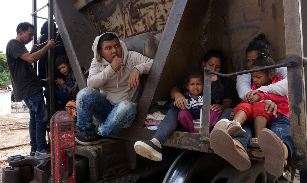 People sit on the side of a freight train car. Two women grip toddlers in their laps.