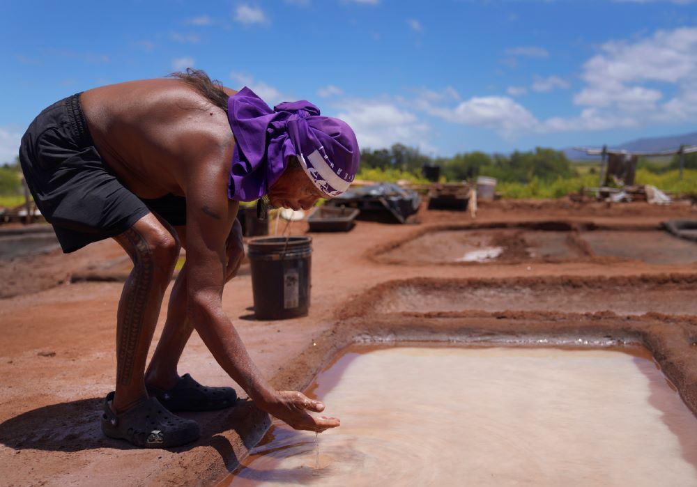 Kane Turalde reaches into a salt bed to examine the salt crystals on Friday, July 14, 2023, in Hanapepe, Hawaii. Climate change, air pollution and littering by tourists and visitors are all threats to this practice, but the 22 families who continue this tradition are fighting to keep these threats at bay and pass on this sacred practice to future generations. (AP Photo/Jessie Wardarski)