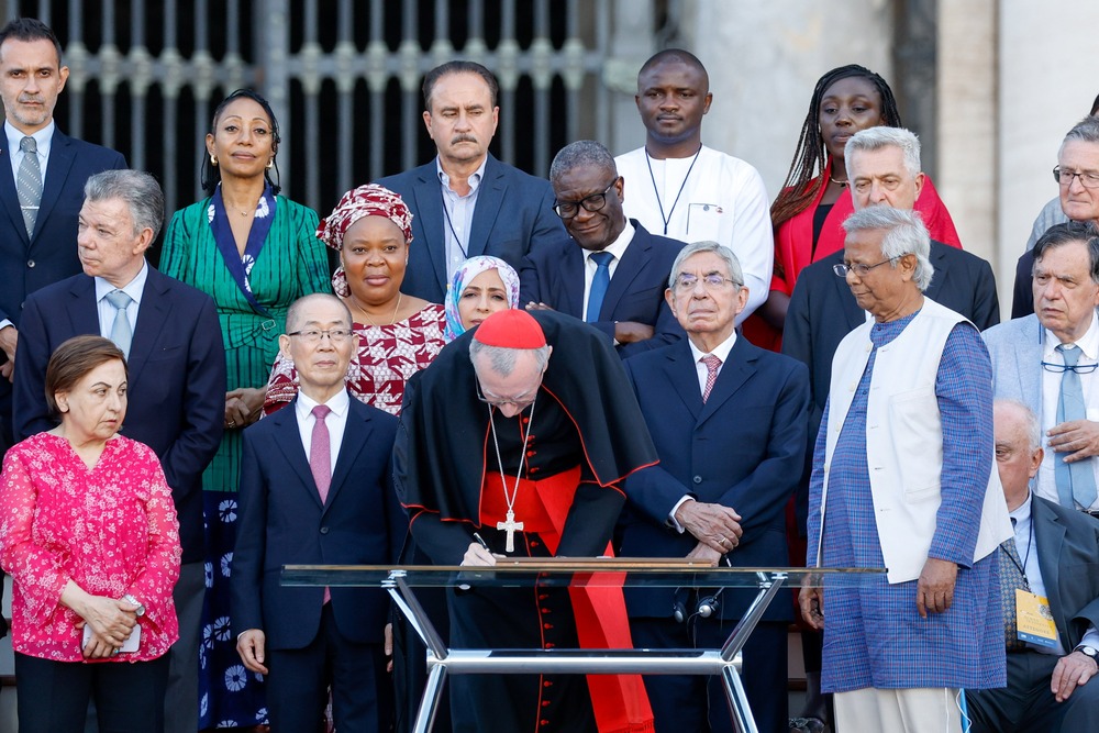 Cardinal bends over table to sign document, with others in background. 