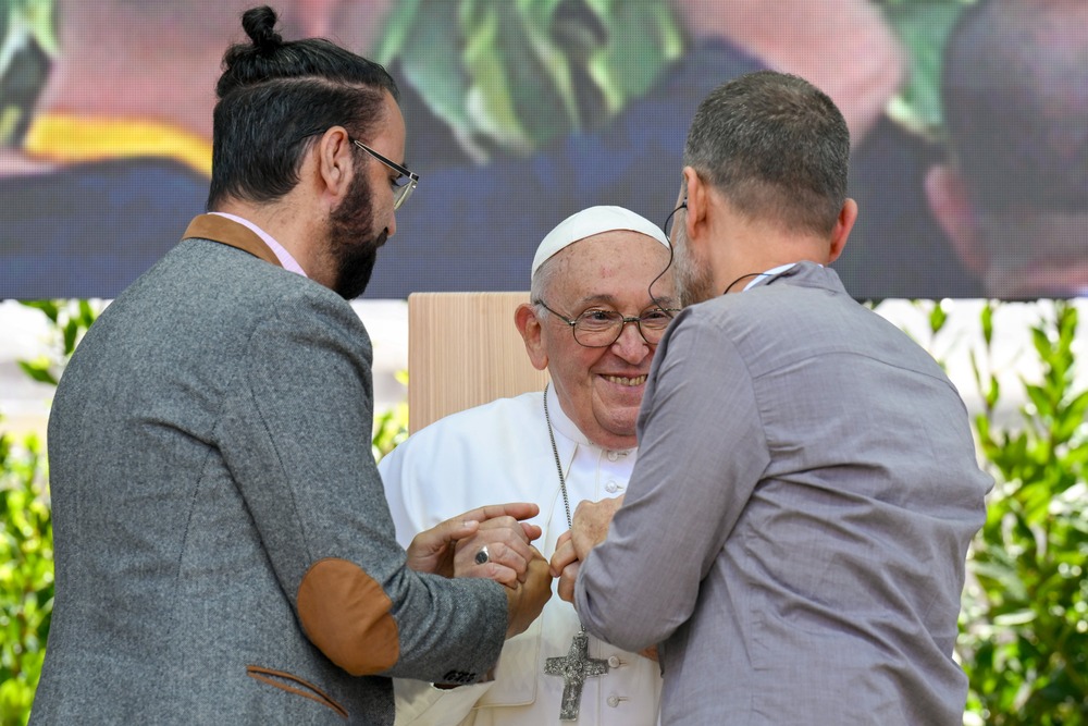 Pope Francis, seated, smiles and takes the hands of the two men. 