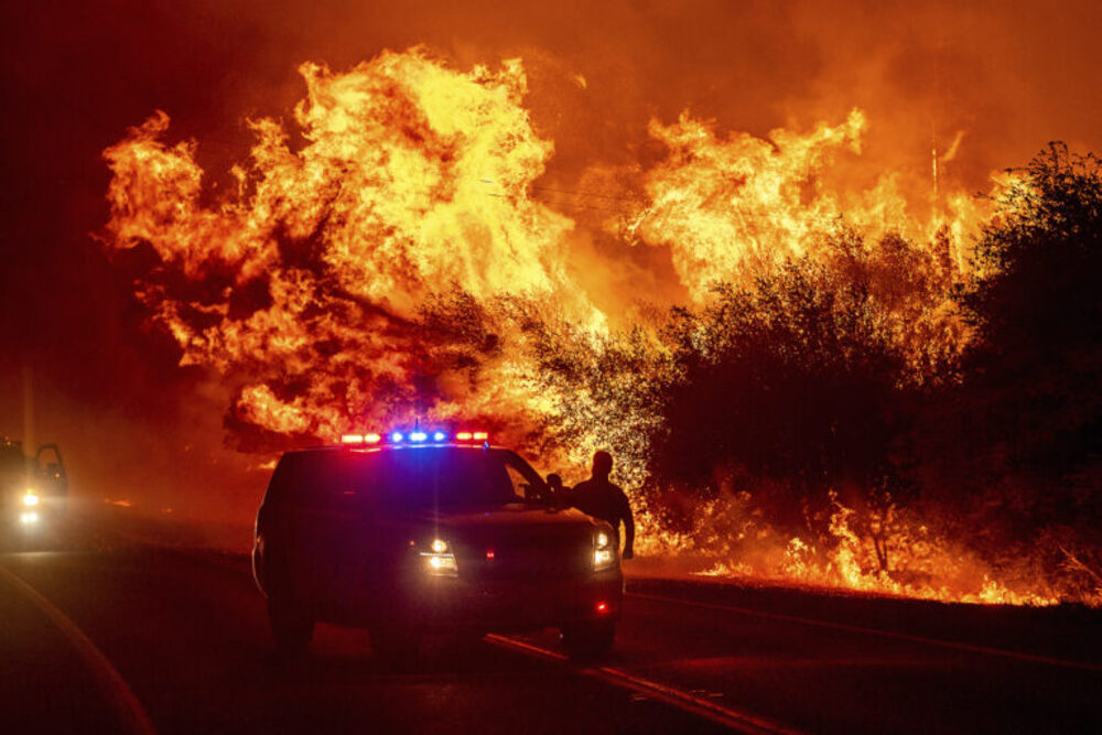 Towering conflagration pierces night sky, emergency vehicle seen in foreground