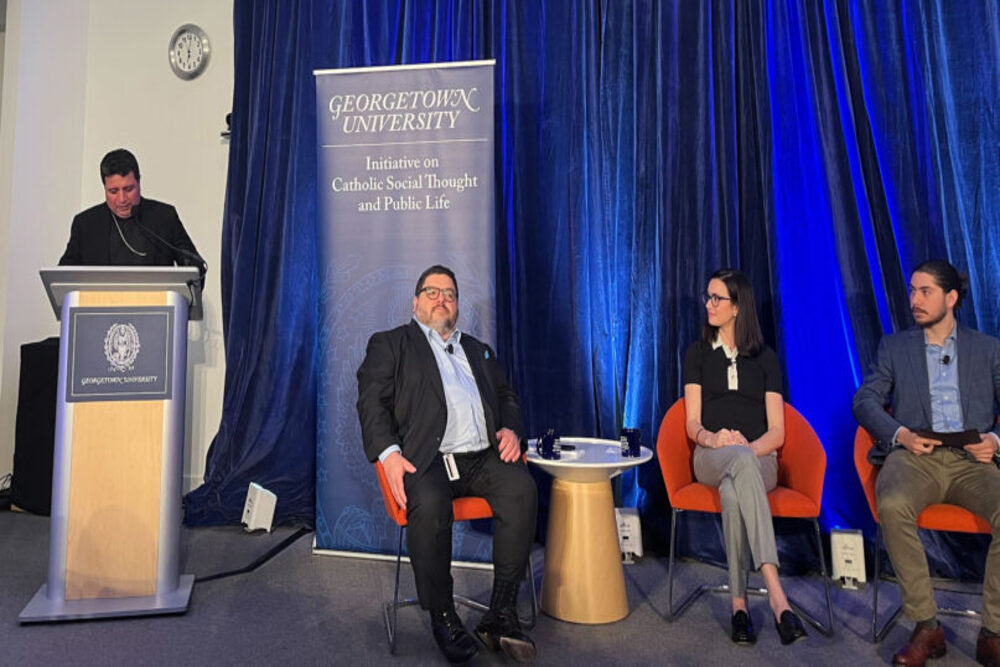 Three members seated, and one standing at lectern in front of blue backdrop