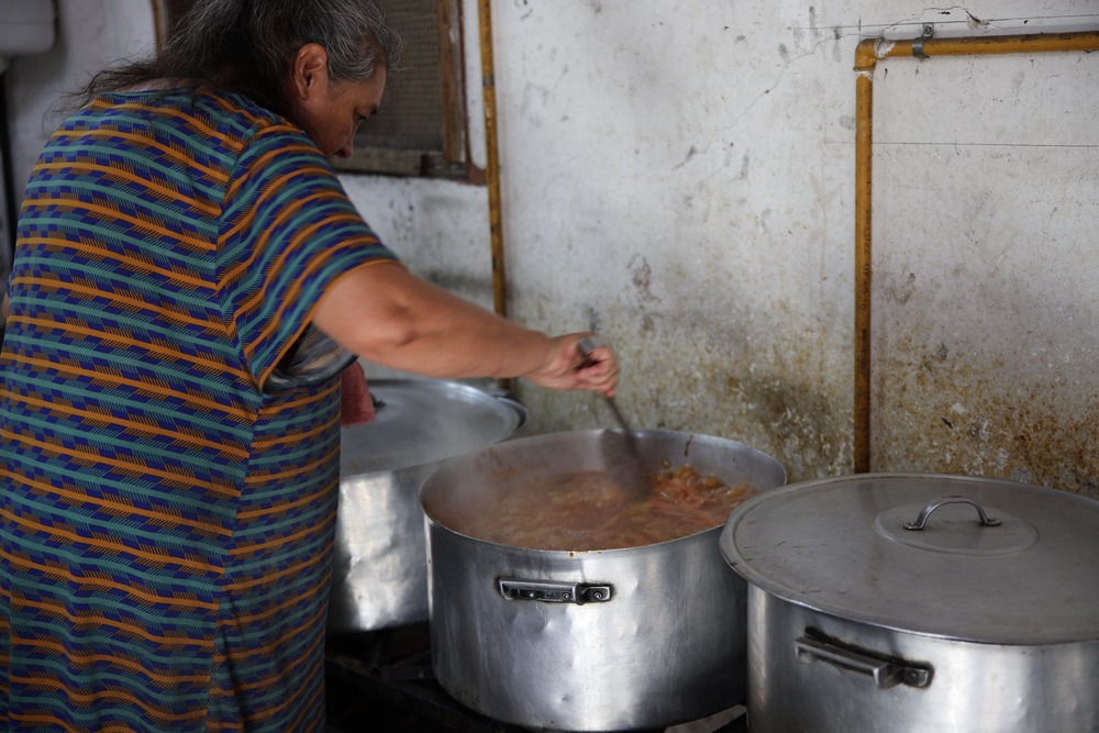 Woman bends over stove stirring food in large aluminum pots