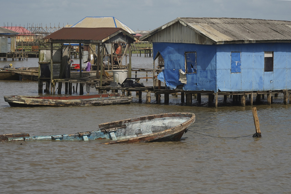 Nigeria coastal erosion In background are houses on stilts, sitting just barely above the water line, and foregrounded is a partially submerged boat tied to dock.