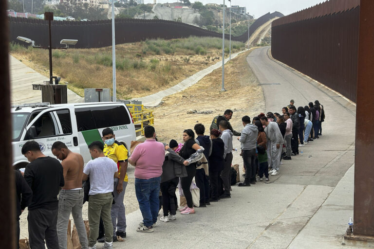 People line along border wall, facing official vehicle