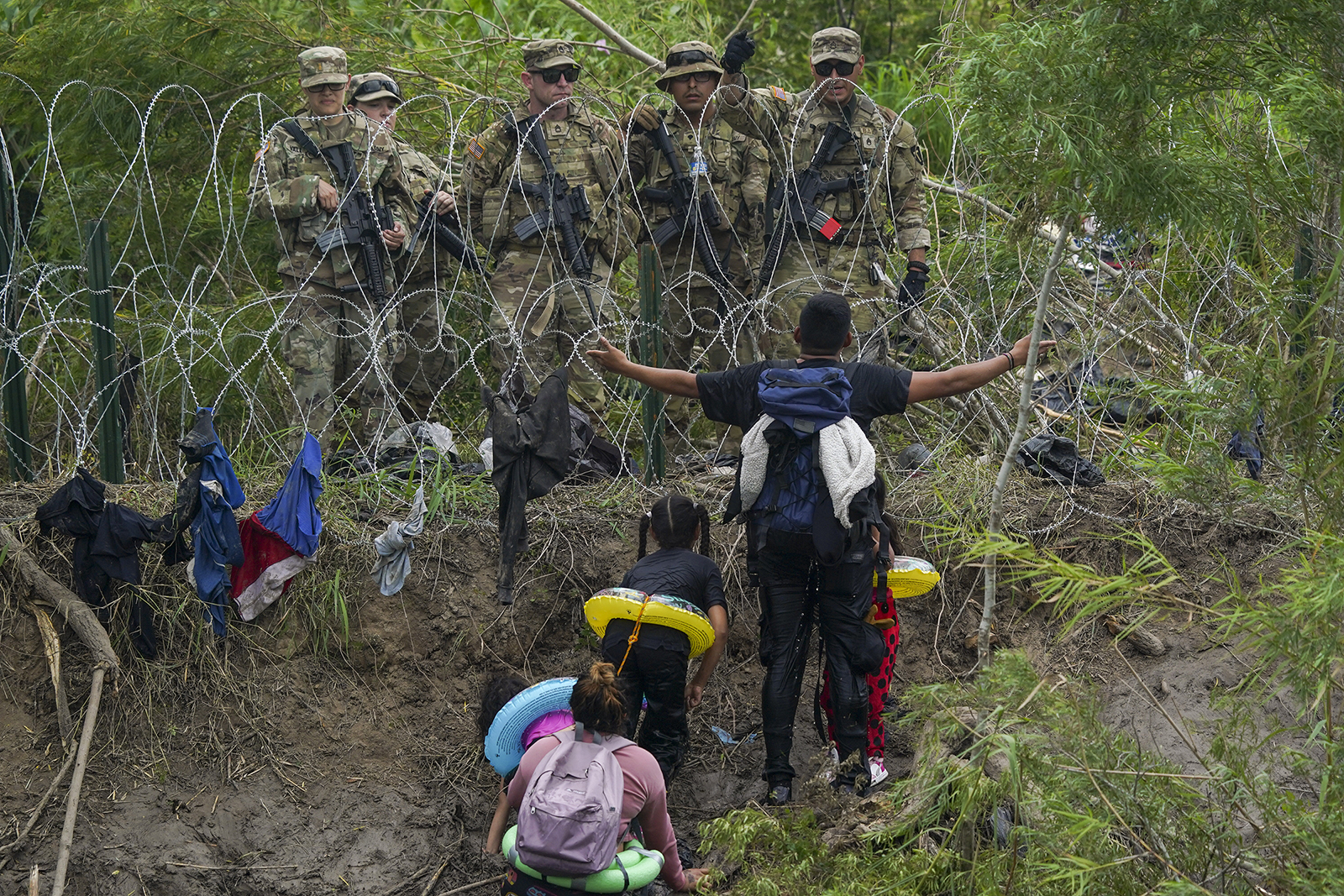 Man stretches arms out shielding his crouching family, while on other side of concertina wire, immigration officers turn him back