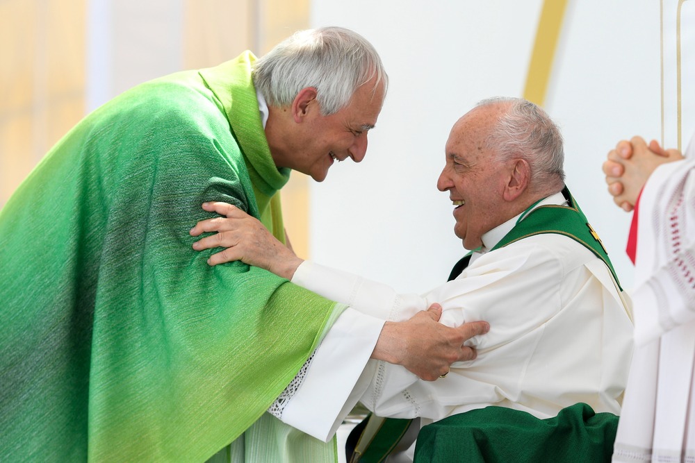 Zuppi, vested, bends to Francis, seated, as the embrace each other.