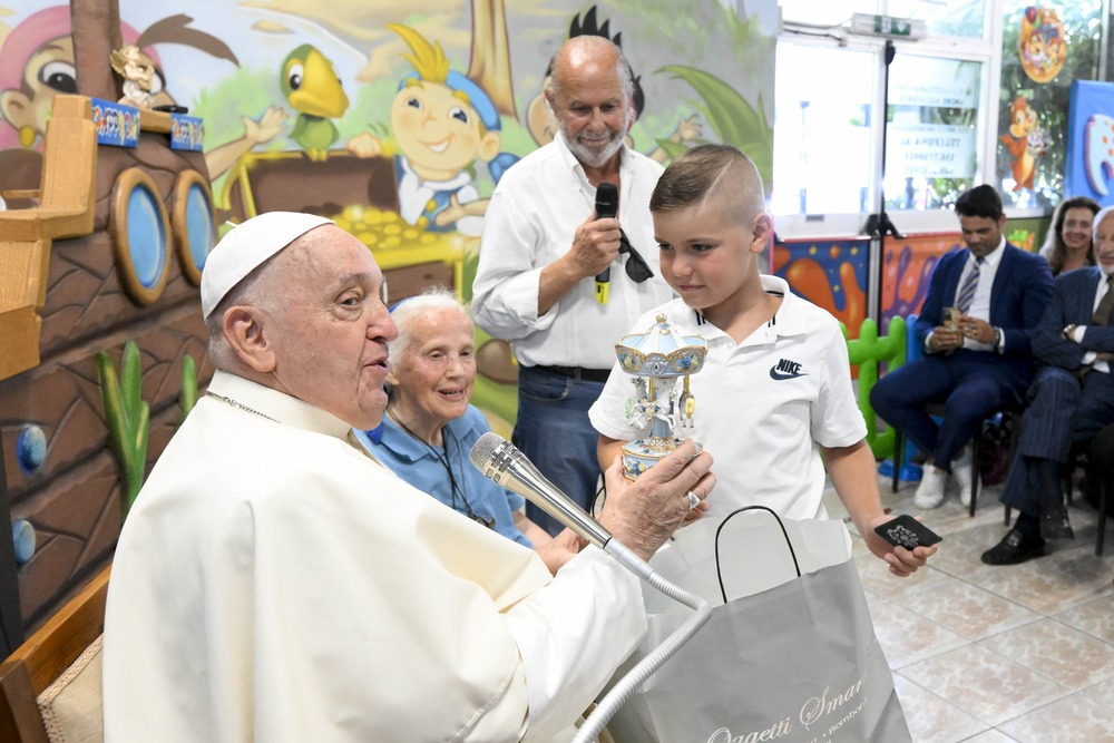 Pope Francis, seated next Sister Jeanningros, holds carousel and smiles; young boy who presented gift stands in front of Francis, watching his reaction. 