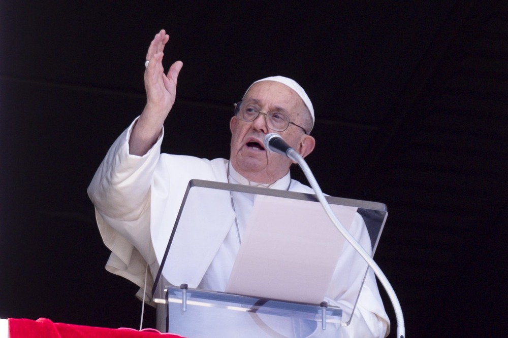 Francis stands in window of Apostolic Palace, in front of lectern, hand raised in blessing. 