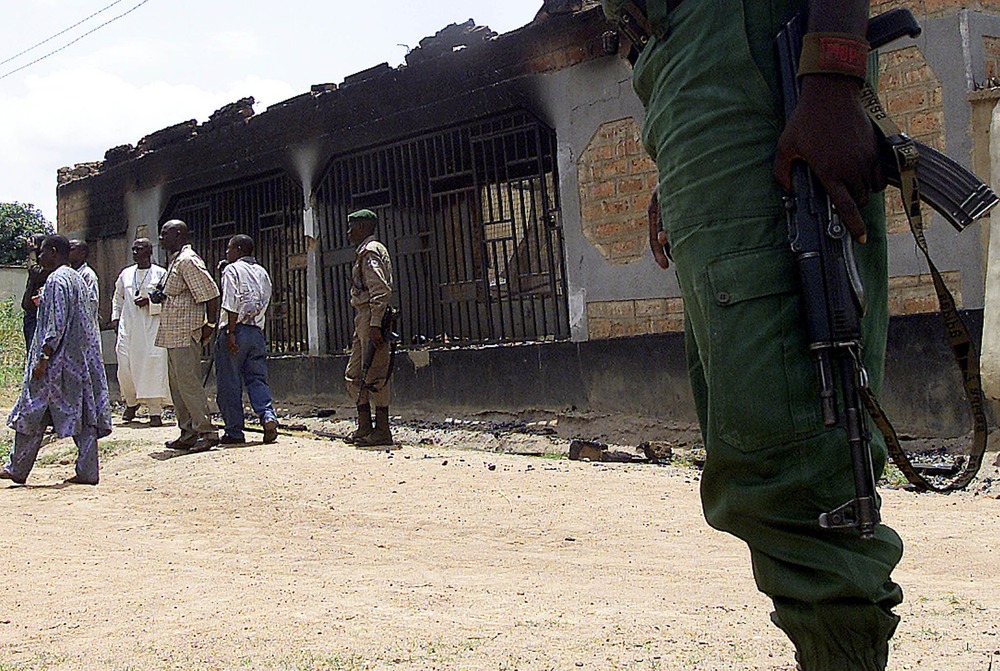 Nigerian security personnel; Adikpo, Nigeria. Armed police stand outside burnt shell of building; civilians come and go.