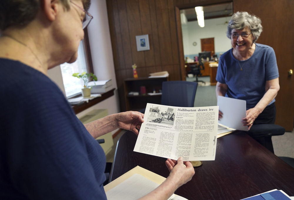 The two sisters, wearing plainclothes, stand in wood-paneled office, looking at clippings and smiling. 