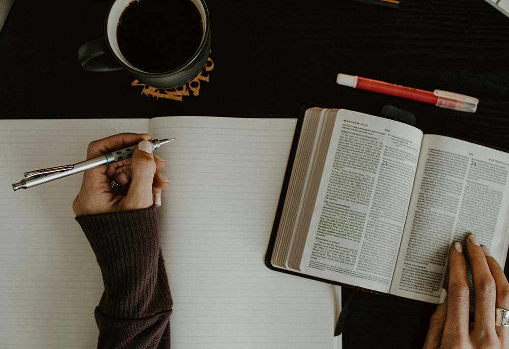 An photo illustration displays an overhead shot of a woman writing in a notebook, referencing a Bible. (Unsplash/Kelly Sikkema)