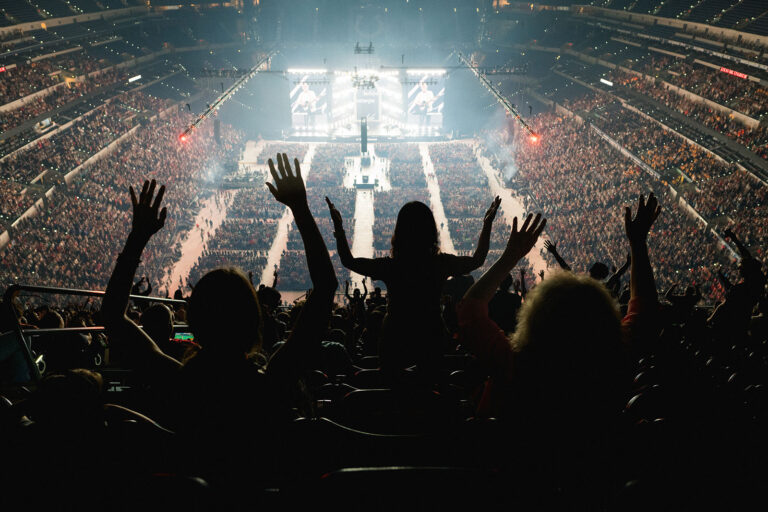 Back view of people in nosebleed seats raising hands; light emanates from distant stage. 