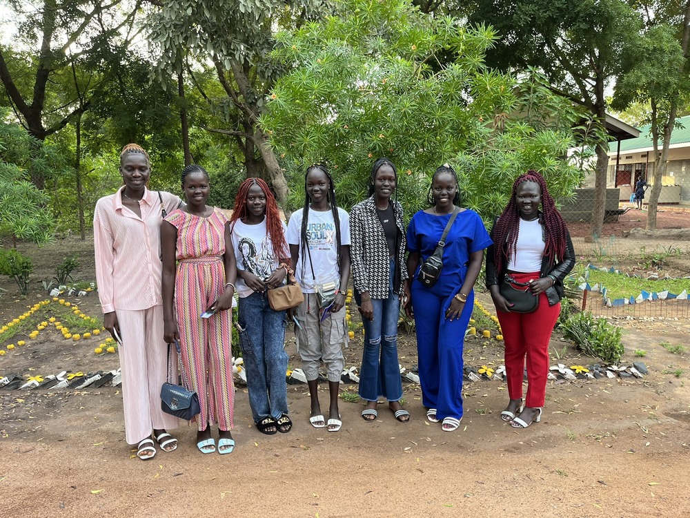 Seven young women stand in  row. In the background are trees, garden beds, and buildings.