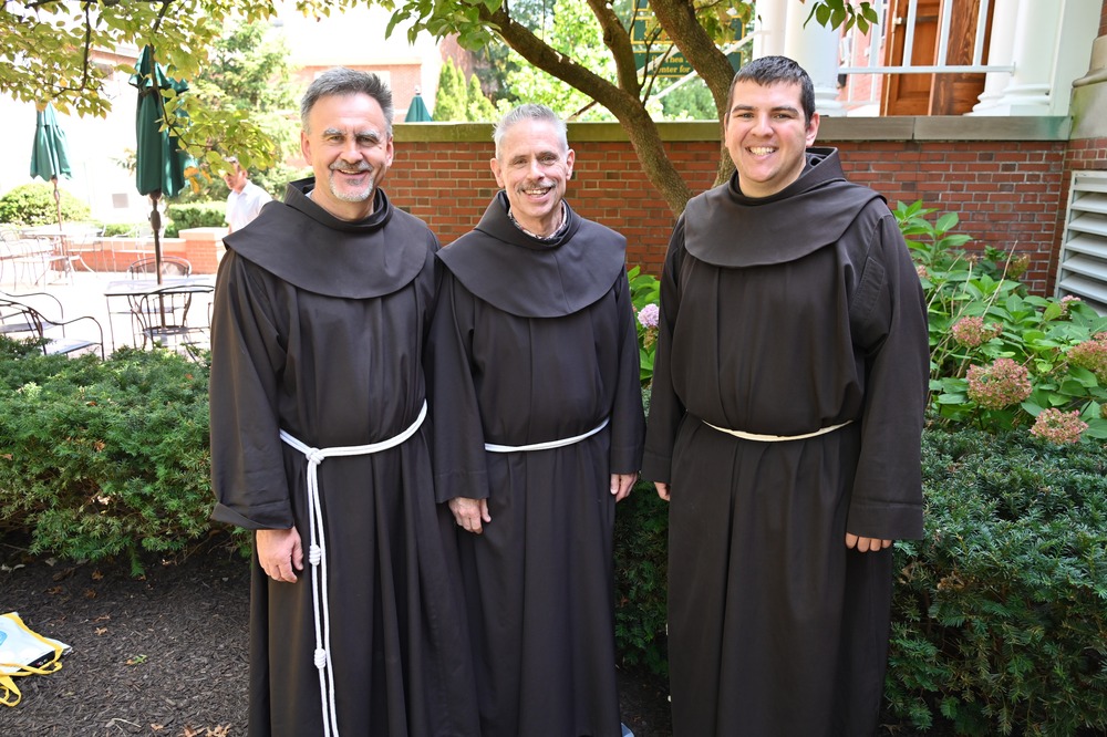 The three friars stand under a tree smiling. 